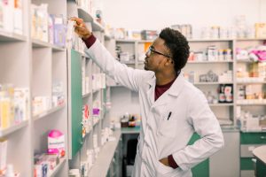 Confident African American man pharmacist standing in interior of pharmacy and searching the drug on shelves in pharmacy